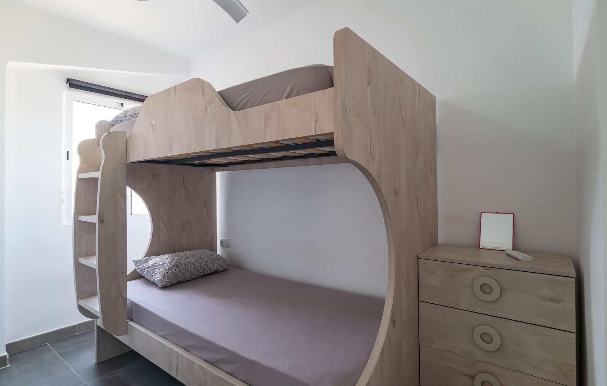 Ein Stockbett mit Holzrahmen, einer Matratze und einem Kopfkissen. Ein Schminktisch mit Schubladen und ein kleiner Tisch mit rotem Gestell sind ebenfalls vorhanden.