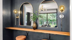 Une pièce avec un mur sombre, un banc en bois, deux tabourets, un vase avec une plante et deux miroirs fixés au mur, équipés de lumières.