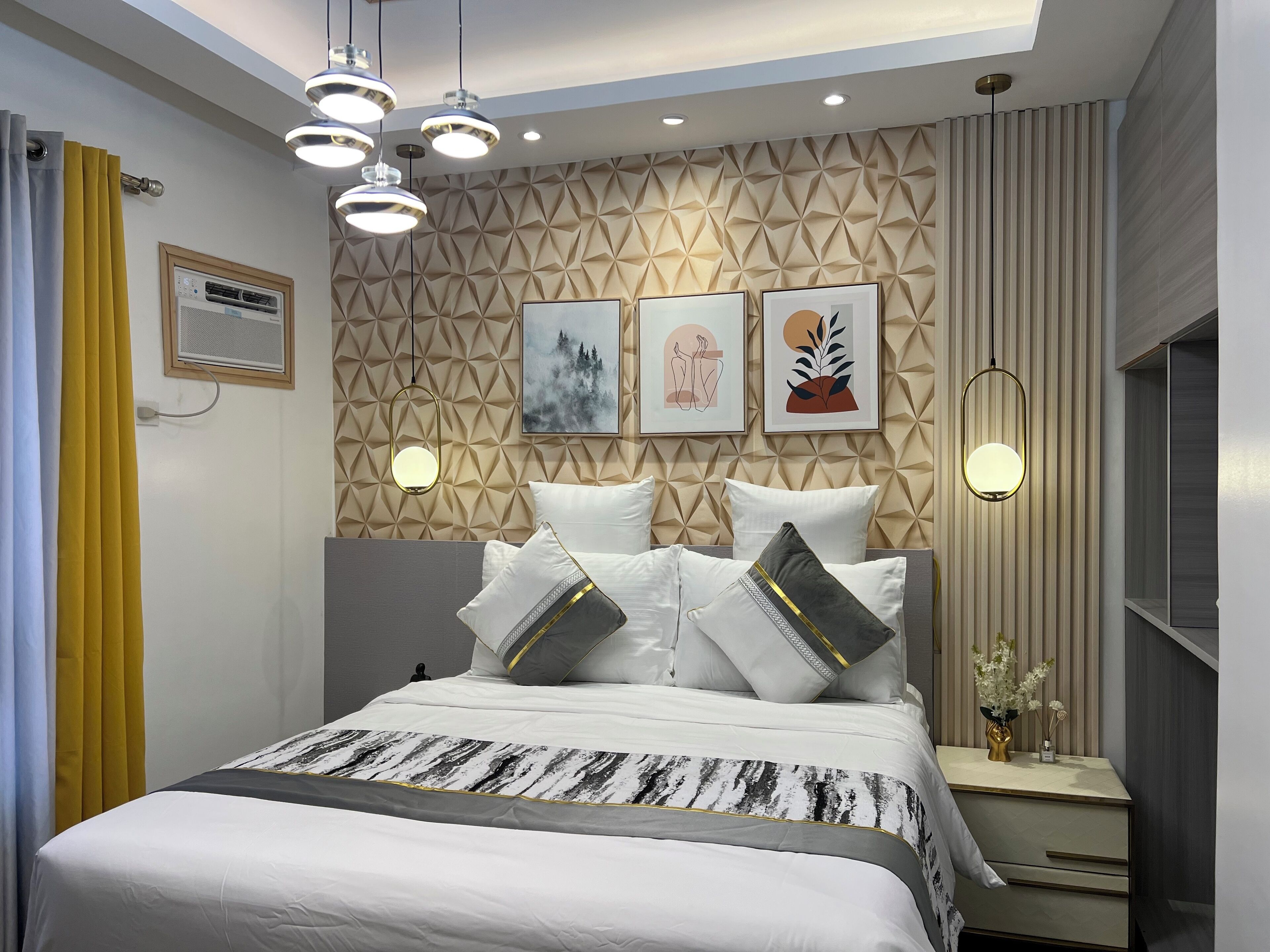 A modern bedroom with a geometric-patterned headboard, a bed with white bedding and decorative pillows, a bedside table with a vase of flowers, and a framed artwork above the bed.