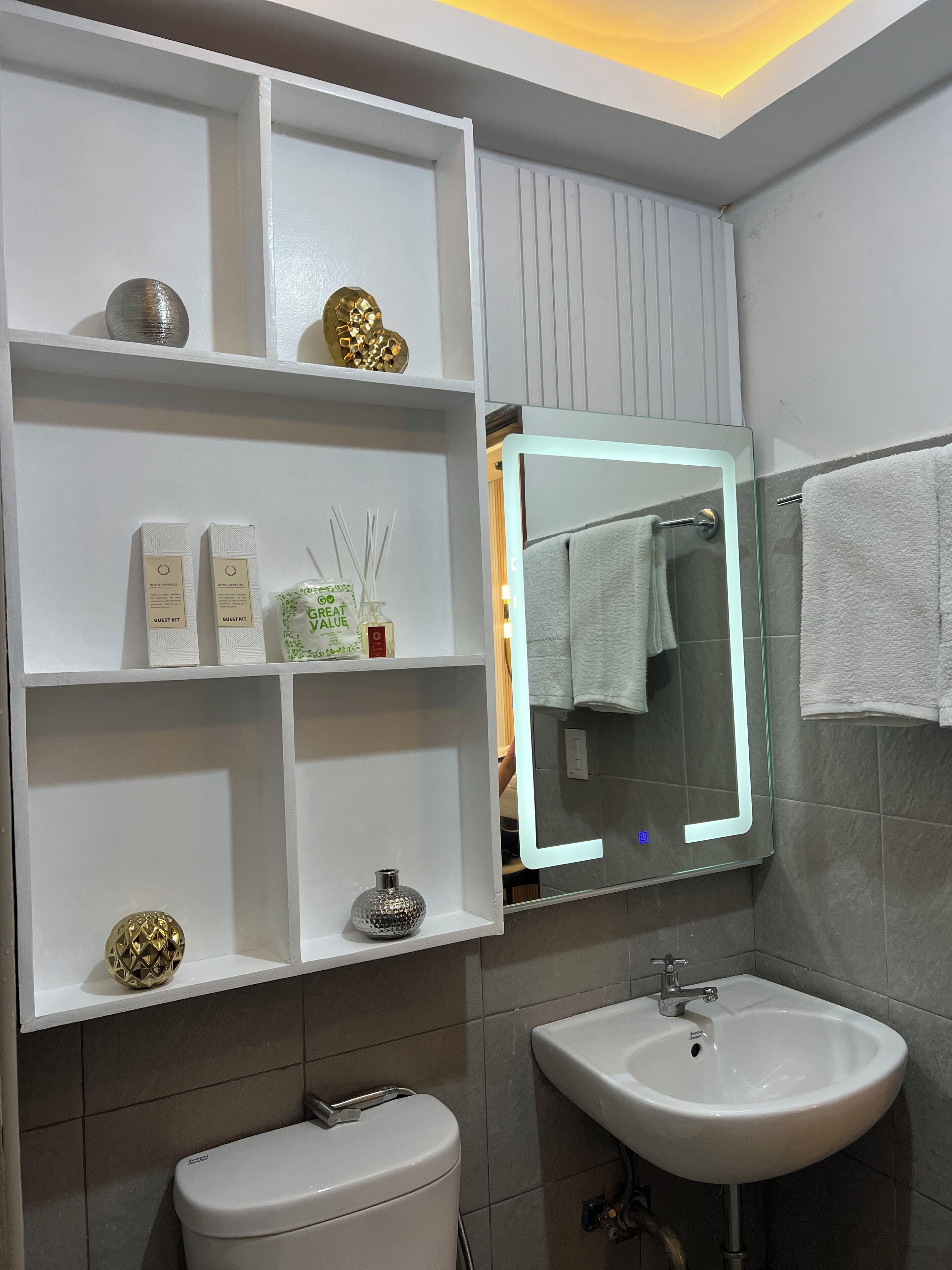 Ein Badezimmer mit einem weißen Waschbecken, einer Toilette und einem Spiegel mit beleuchtetem Rahmen. Regale mit Pflegeprodukten und Dekorationsobjekten sind vorhanden.