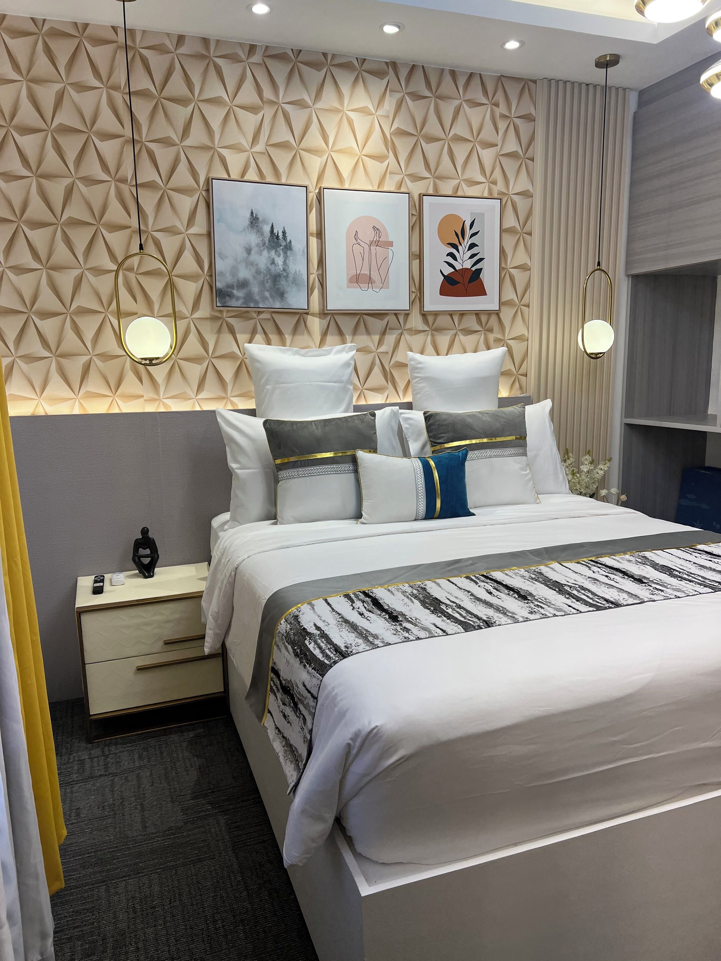 A modern bedroom with a geometric-patterned headboard, a neatly made bed with multiple pillows, bedside tables with lamps, and framed artwork on the wall.