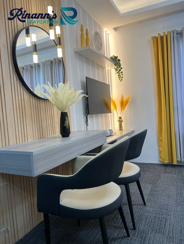 Una habitación de hotel moderna con un escritorio de madera, dos sillas negras, un espejo redondo, un televisor de pantalla plana y una planta en maceta.