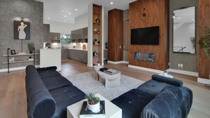 Una sala moderna con sofás oscuros, una mesa de centro con un libro y una planta, un televisor montado en la pared y una cocina al fondo.
