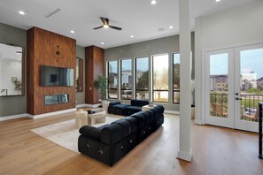 A spacious living area with a large sectional sofa, a flat-screen TV mounted on a wooden wall, and a balcony with a view of the city. - Newly Constructed Winsome Village B - Galleria Area! (Houston)