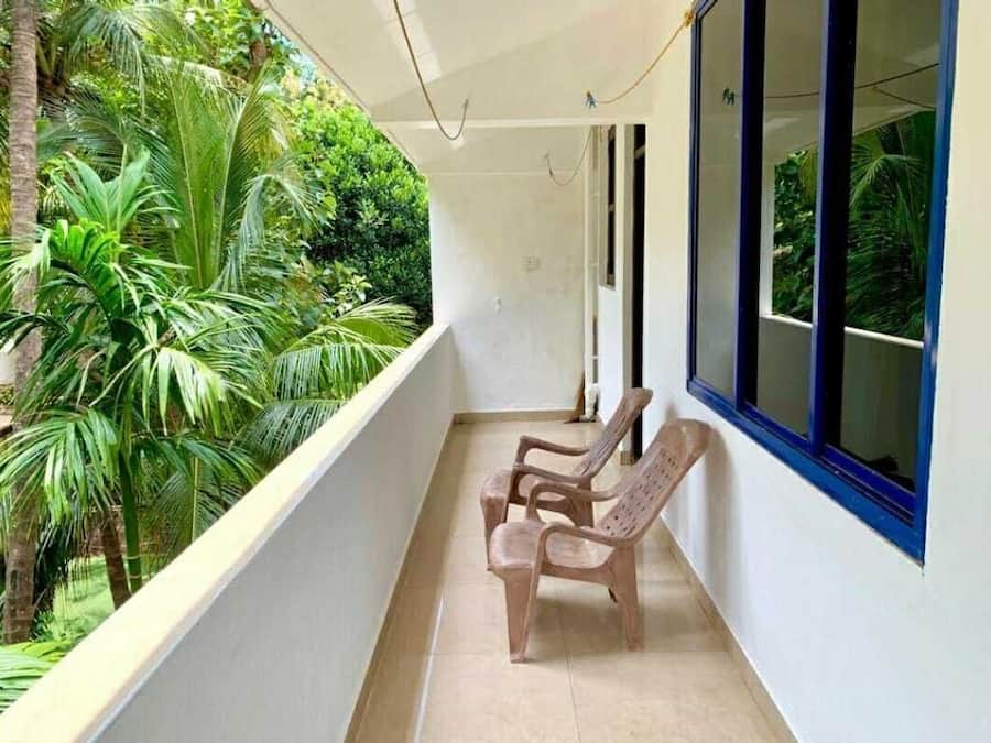 Un balcón con dos sillas de plástico, una pared blanca y una ventana que da a zonas verdes.