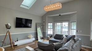 A modern living room with a grey sofa, a flat-screen TV mounted on the wall, and a large window providing natural light.