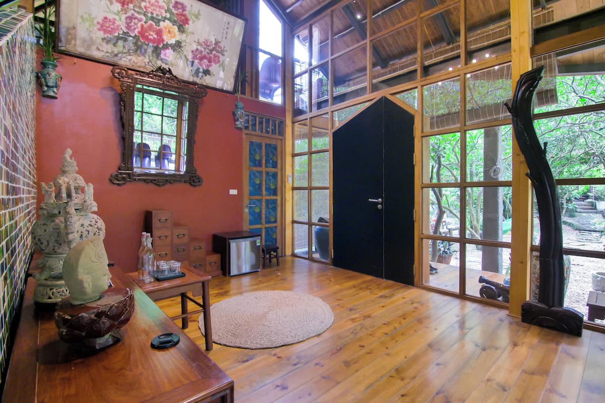 Une pièce spacieuse avec un plancher en bois, une grande fenêtre donnant sur un paysage verdoyant, une porte noire et une petite table agrémentée d’objets décoratifs.