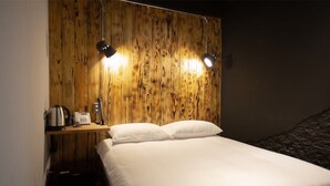 A room with a wooden headboard, two bedside lamps, a bed with white linens, a small table with a kettle and a phone, and a dark wall.