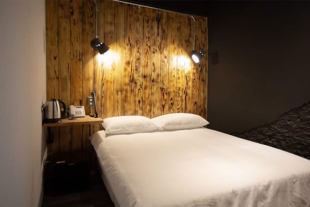 Une chambre avec une tête de lit en bois, deux lampes de chevet, un lit avec du linge de lit blanc, une petite table avec une bouilloire et un téléphone, et un mur de couleur sombre.