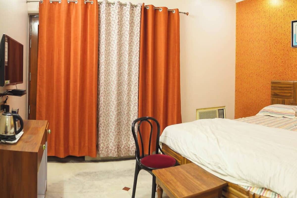 A hotel room with a bed, a desk with a kettle, a chair, and a picture on the wall.