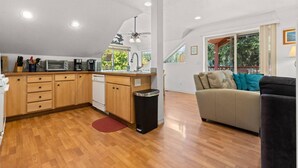 A modern kitchen with wooden cabinets, a dishwasher, and a sink. There is a dining area with a sofa and a window offering a view of greenery. - Casa Twin Lakes | Private Dock, Lake View, Fire Pit, Large Yard, Near Silverwood (Rathdrum)