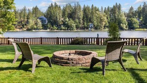A grassy outdoor area with wicker chairs and a fire pit, overlooking a lake and forested area. - Casa Twin Lakes | Private Dock, Lake View, Fire Pit, Large Yard, Near Silverwood (Rathdrum)