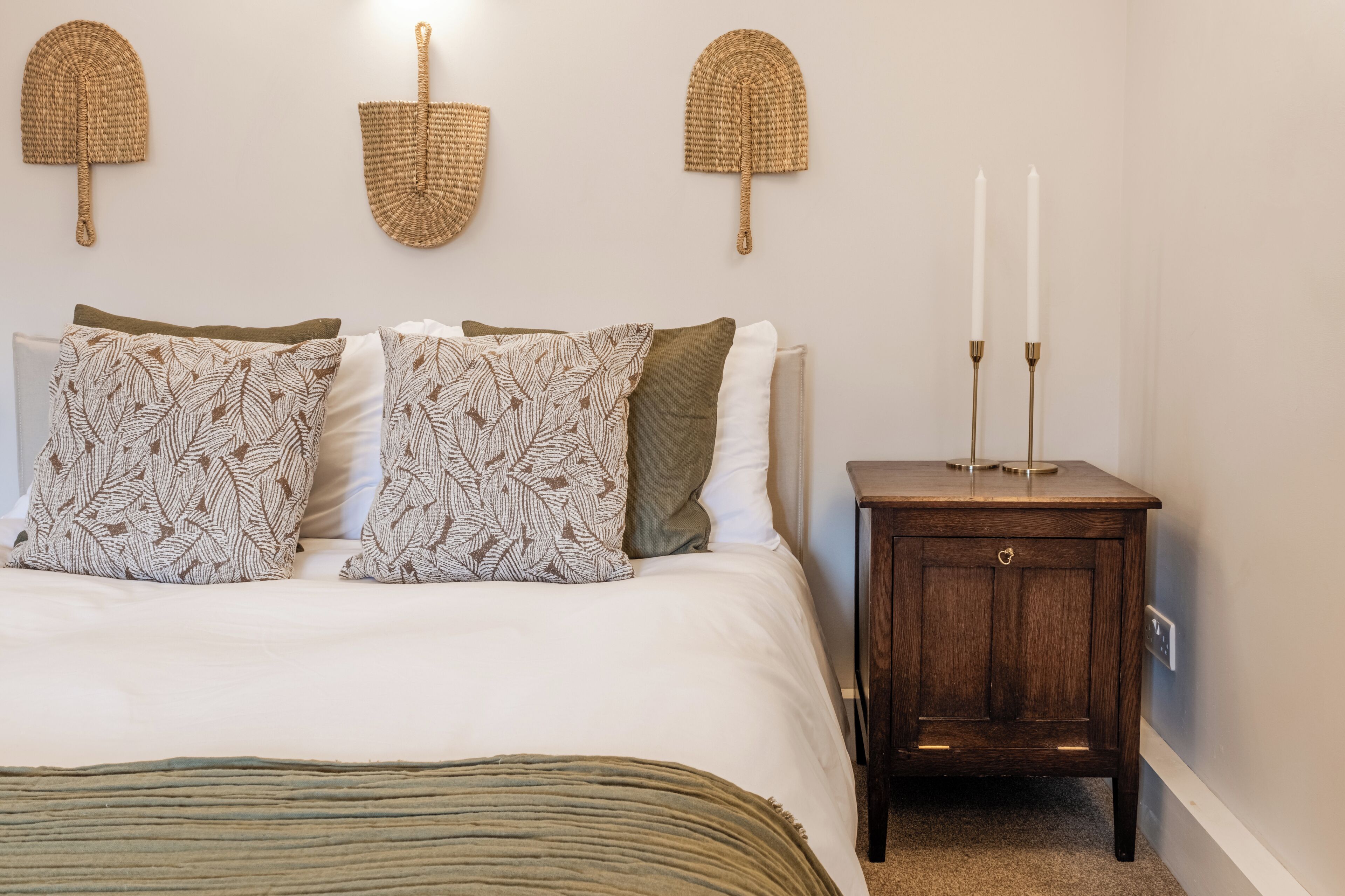 A neatly made bed with patterned pillows, a dark wooden nightstand with two candles, and woven wall hangings.