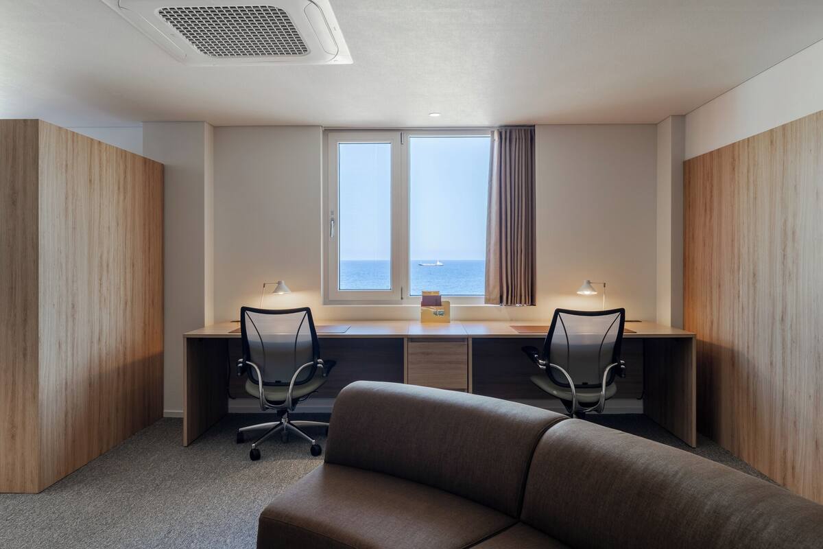 A modern hotel room with a large desk, two chairs, a sofa, and a large window with a view of the sea.