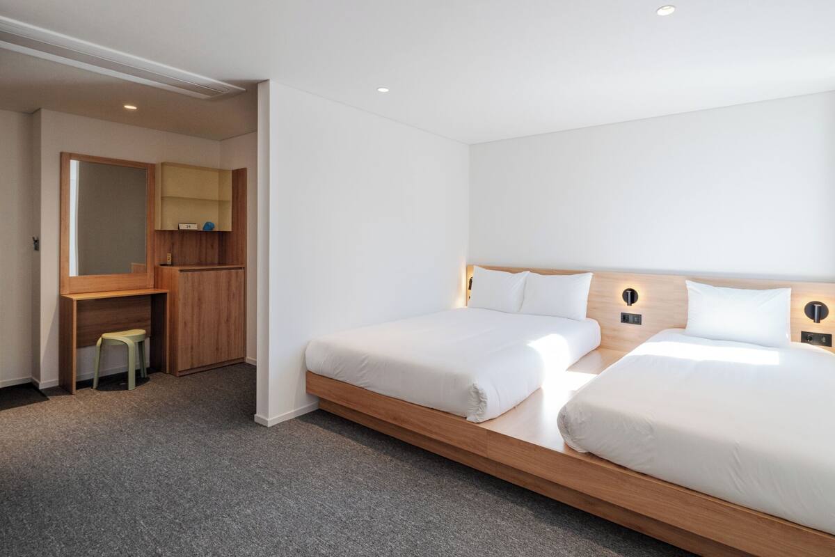 A modern hotel room with two beds, a wooden desk, and a small stool.