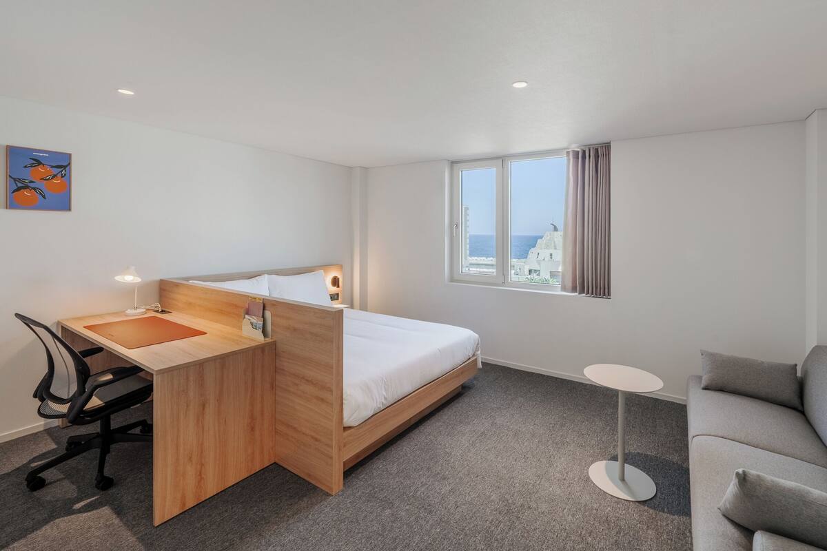 A hotel room with a bed, a desk with a chair, a small round table, and a view of the sea.