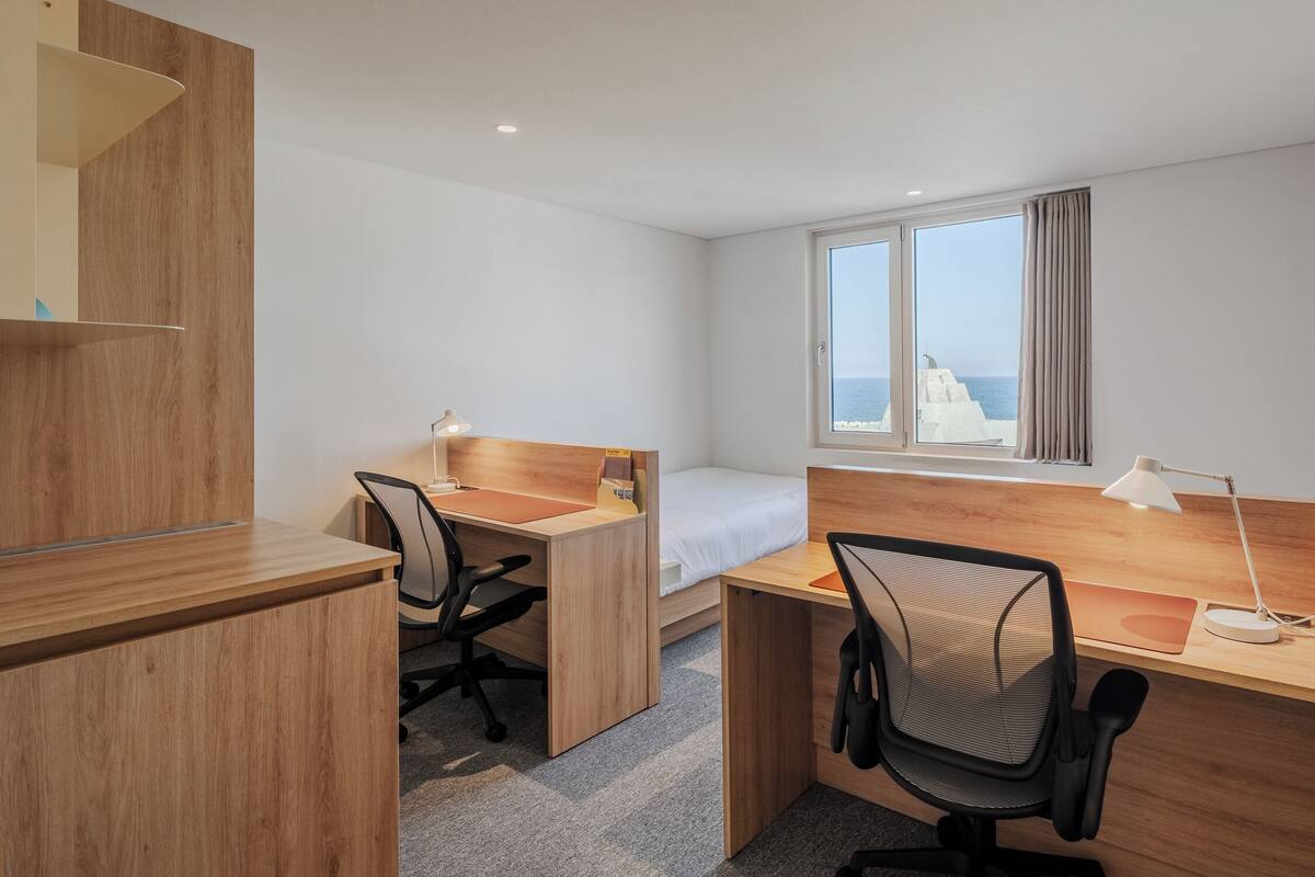 A hotel room with a wooden desk, two chairs, a bed, and a window with curtains.