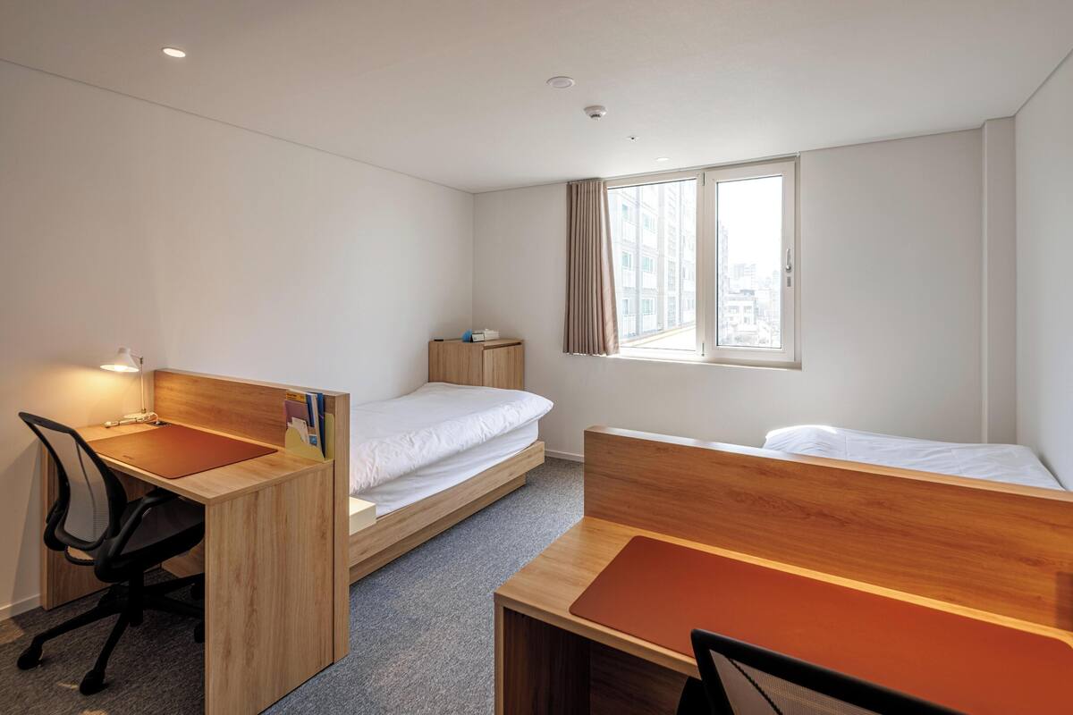 A compact hotel room with a single bed, a desk with a chair, and a window providing a view of the city.