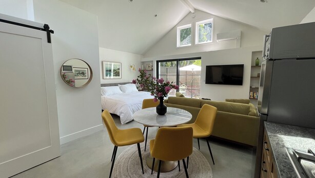 A modern hotel room with a bed, a dining area with yellow chairs, a sofa, and a kitchen area with a refrigerator.