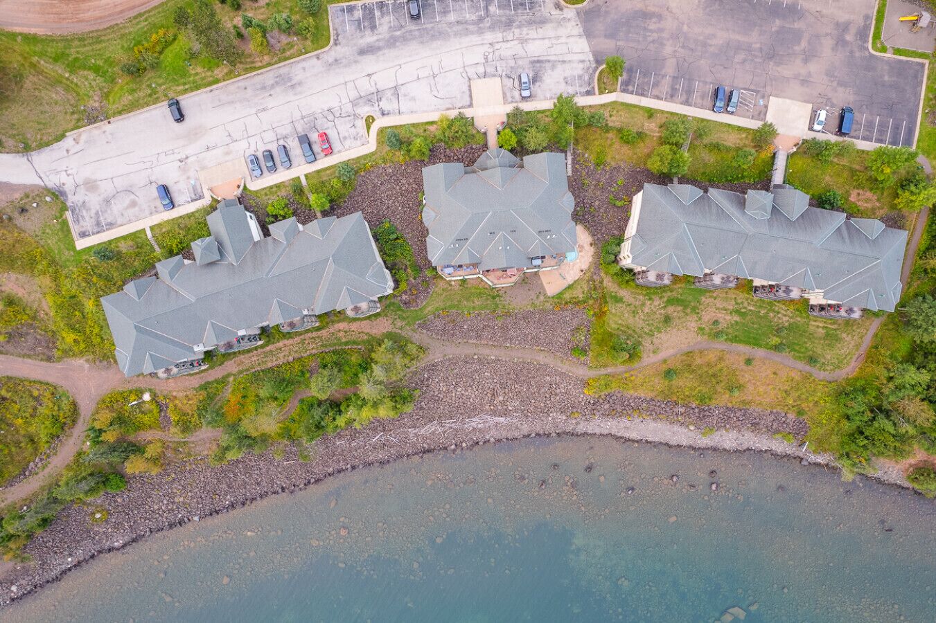 An aerial view of a coastal property with three buildings, a parking lot, and a rocky shoreline.