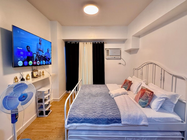 Habitación de hotel con una cama, televisor montado en la pared, ventilador de pie y radiador.