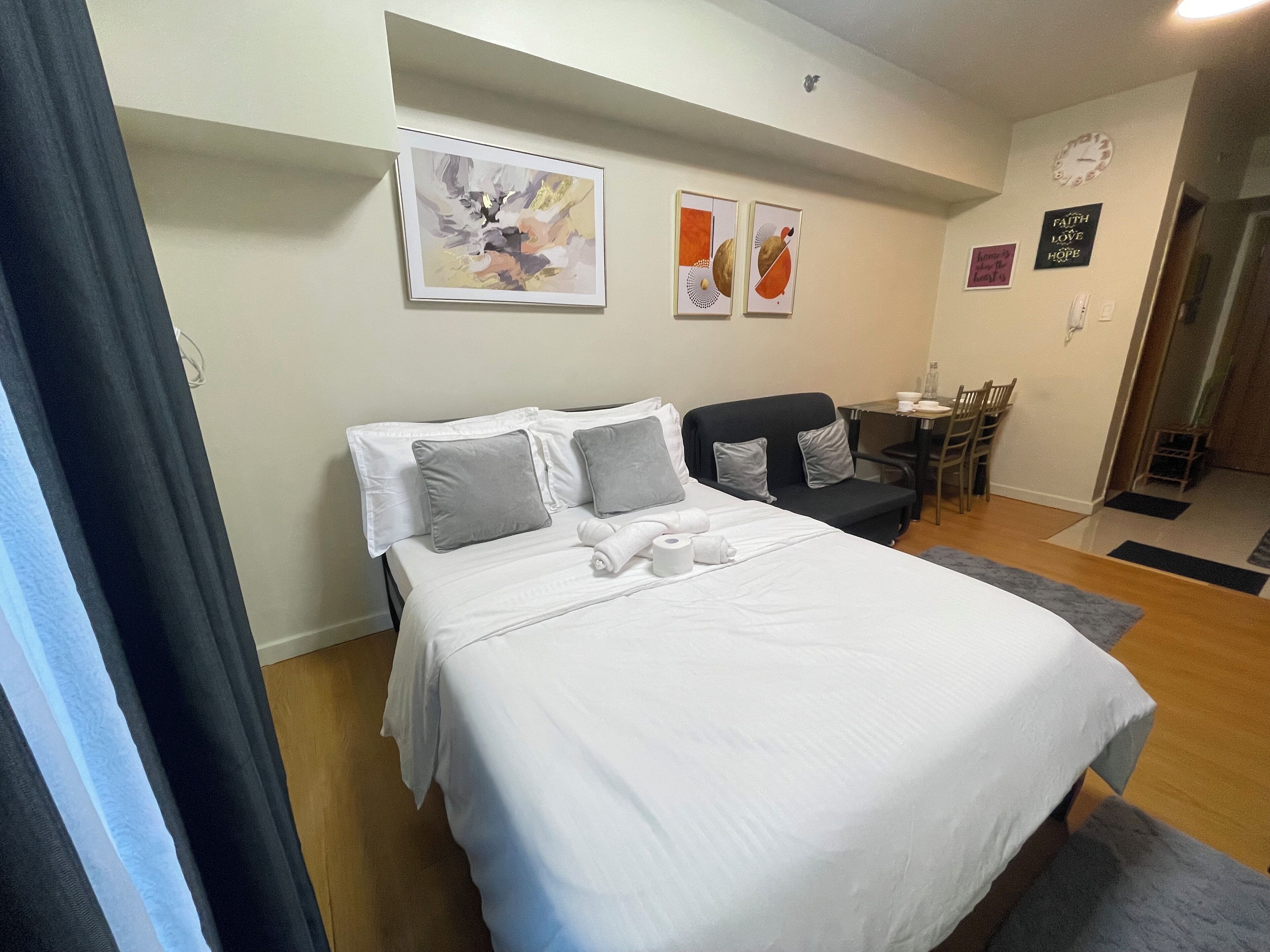 A neatly made bed with white linens and grey pillows, a small black sofa, a dining table with chairs, and a wall clock.