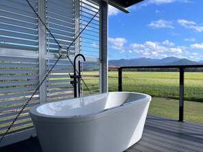 Une baignoire blanche, installée en extérieur sur une terrasse en bois, offre une vue sur un champ verdoyant et des montagnes en arrière-plan.
