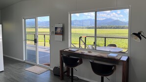 A modern room with a large window offering a view of a green field and mountains. There is a desk with two chairs and a lamp. - Pinnacolo Farm Stay (Pinnacle)