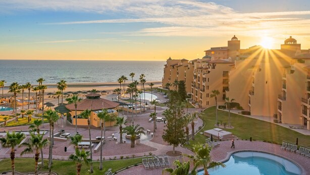 A resort with a pool, palm trees, and a view of the ocean at sunset.