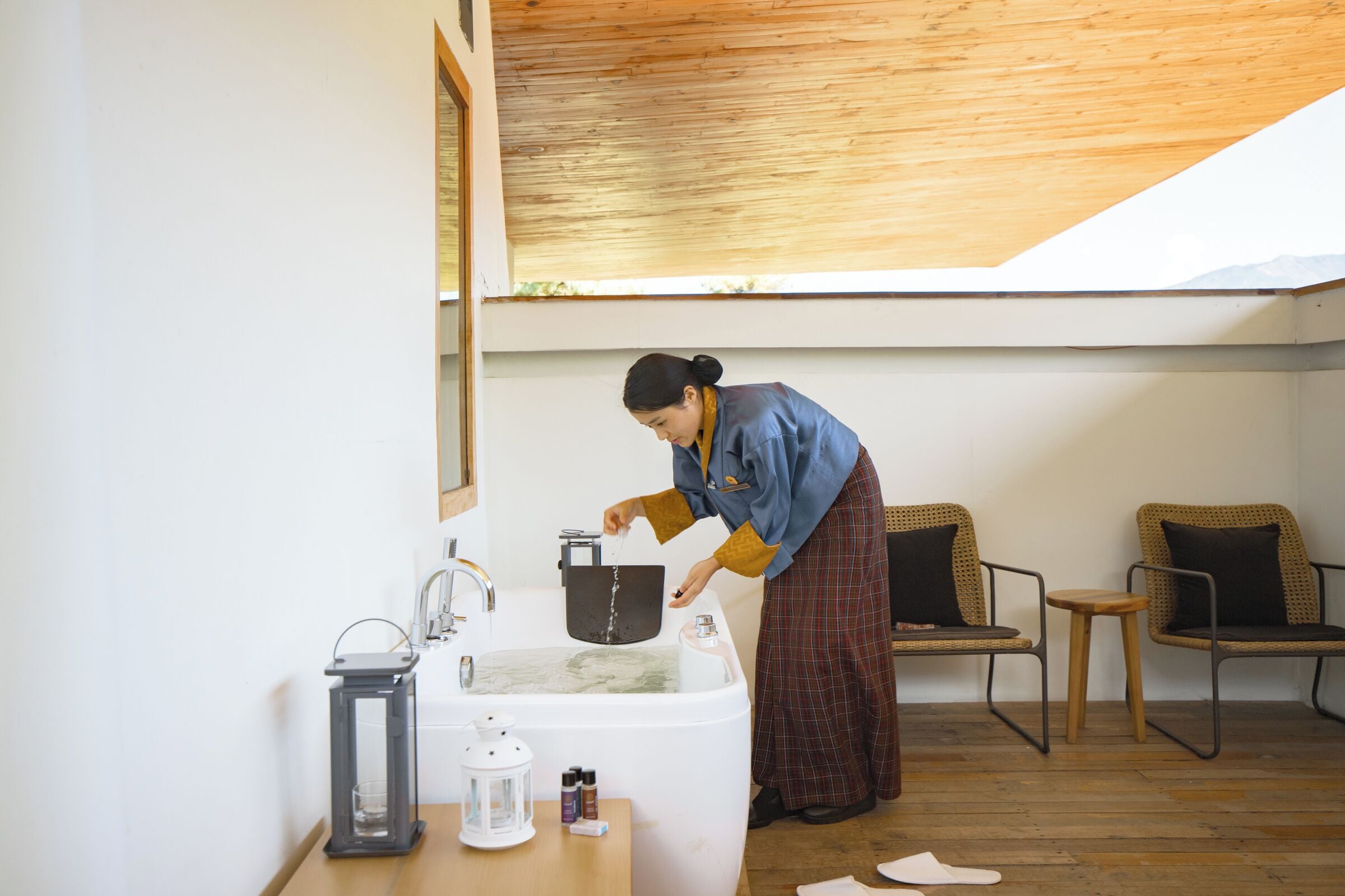 A person washing dishes in a modern kitchen with a wooden ceiling, a white sink, and a small table with a lantern and condiments.