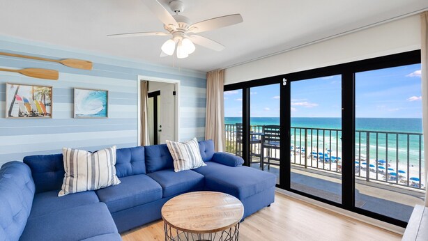 Un salon spacieux avec un canapé bleu, une table basse en bois et un ventilateur de plafond. Une grande fenêtre offre une vue sur la plage et l’océan.