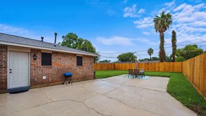 A backyard with a brick house, a wooden fence, a patio with chairs, and a blue grill. - Cozy Coastal Stay: Near Kemah + NASA | BBQ | Beach (Seabrook)