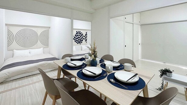A modern dining area with a wooden table set for four, surrounded by chairs. There is a bed with white bedding and a headboard with circular patterns. The room has white walls and a large window.