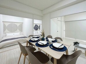 A modern dining area with a wooden table set for four, surrounded by chairs. There is a bed with white bedding and a headboard with circular patterns. The room has white walls and a large window.