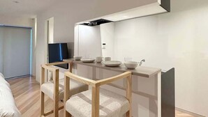 Ein modernes, minimalistisches Zimmer mit einem Holzespeisetisch, zwei Stühlen, einer integrierten Küche inklusive Mikrowelle und einem großen Fenster.