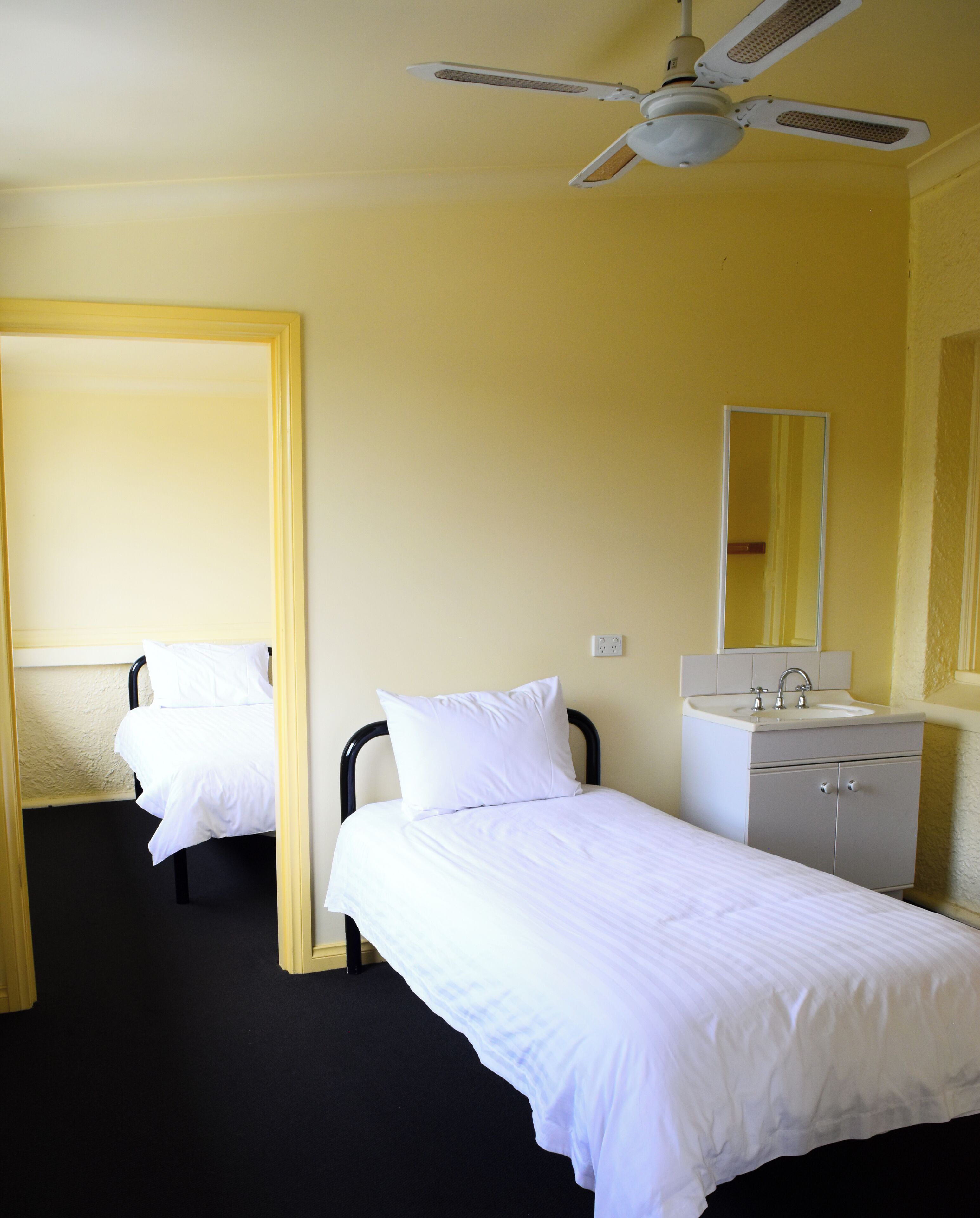 Une chambre d’hôtel avec deux lits simples, un ventilateur de plafond, une salle de bain avec un lavabo et un miroir, et une porte donnant sur une autre chambre.