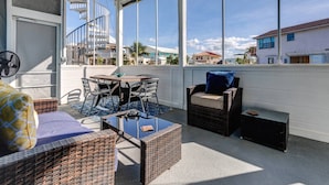 Une terrasse ensoleillée, meublée de meubles en osier, avec une table à plateau de verre et un escalier en colimaçon visible à travers les fenêtres.