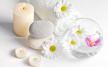 A spa setting with a lit candle, white towels, stones, and a glass bowl with flowers.