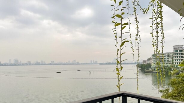 A view of a river with a city skyline in the background, a balcony railing, and hanging plants. - NINE HOUSING 30 Yen Hoa (Hanoi)