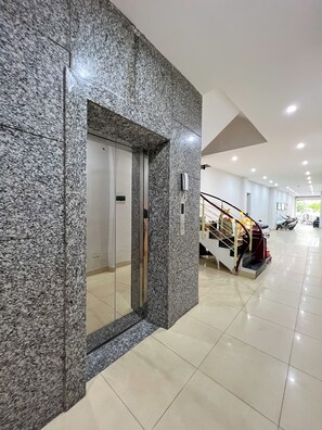 Un intérieur moderne avec un mur en granit, un ascenseur en verre, un escalier doté d’une rampe et un scooter garé au fond.