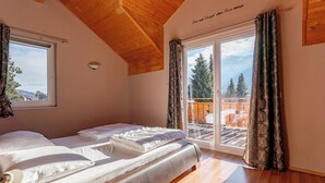 Een slaapkamer met een houten plafond, een bed met witte lakens, een groot raam met gordijnen en uitzicht op een terras en bomen.