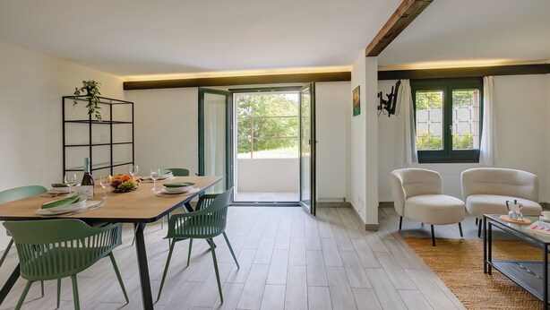 A dining area with a wooden table set for four, green chairs, a plant on a metal shelf, and a view through a glass door to the outside. - Casa Al Cervo (Lugano)