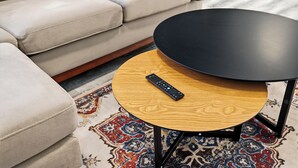 Un canapé beige agrémenté d’un coussin à motifs, une table ronde en bois avec un plateau noir, et une télécommande posée dessus.
