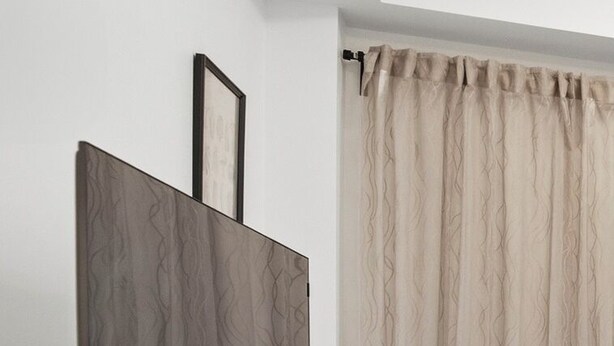 Une pièce avec un meuble TV en bois, un téléviseur à écran plat, un tableau encadré au mur et une fenêtre avec des rideaux beiges.