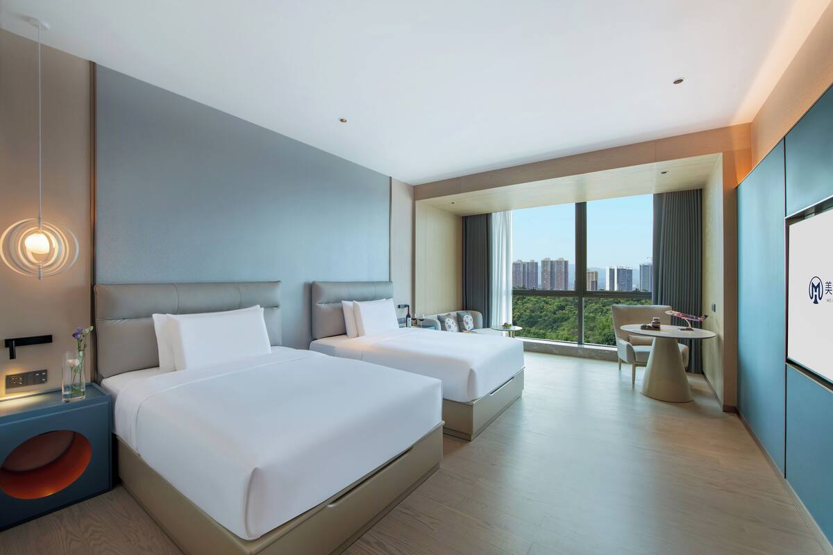 Een moderne hotelkamer met twee bedden, een balkon met uitzicht op de stad, een klein zithoekje en een groot raam.