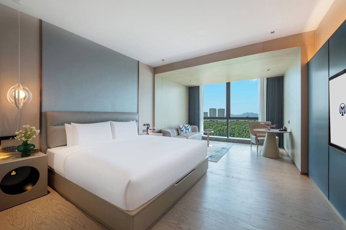 Een moderne hotelkamer met een groot bed, een nachtkastje met lamp, een kleintje tafeltje met stoel, en uitzicht op de stad door grote ramen.