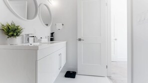Un baño moderno con un tocador blanco, dos espejos, una planta en maceta y un bolso negro en el suelo.