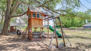 Une cabane en bois, équipée d’un toboggan et de balançoires, entourée d’arbres et donnant sur un espace ouvert.