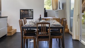 Un espace de repas moderne avec une table dressée pour six personnes, des chaises en bois, un téléviseur fixé au mur et une vue sur l’extérieur grâce à de grandes fenêtres.