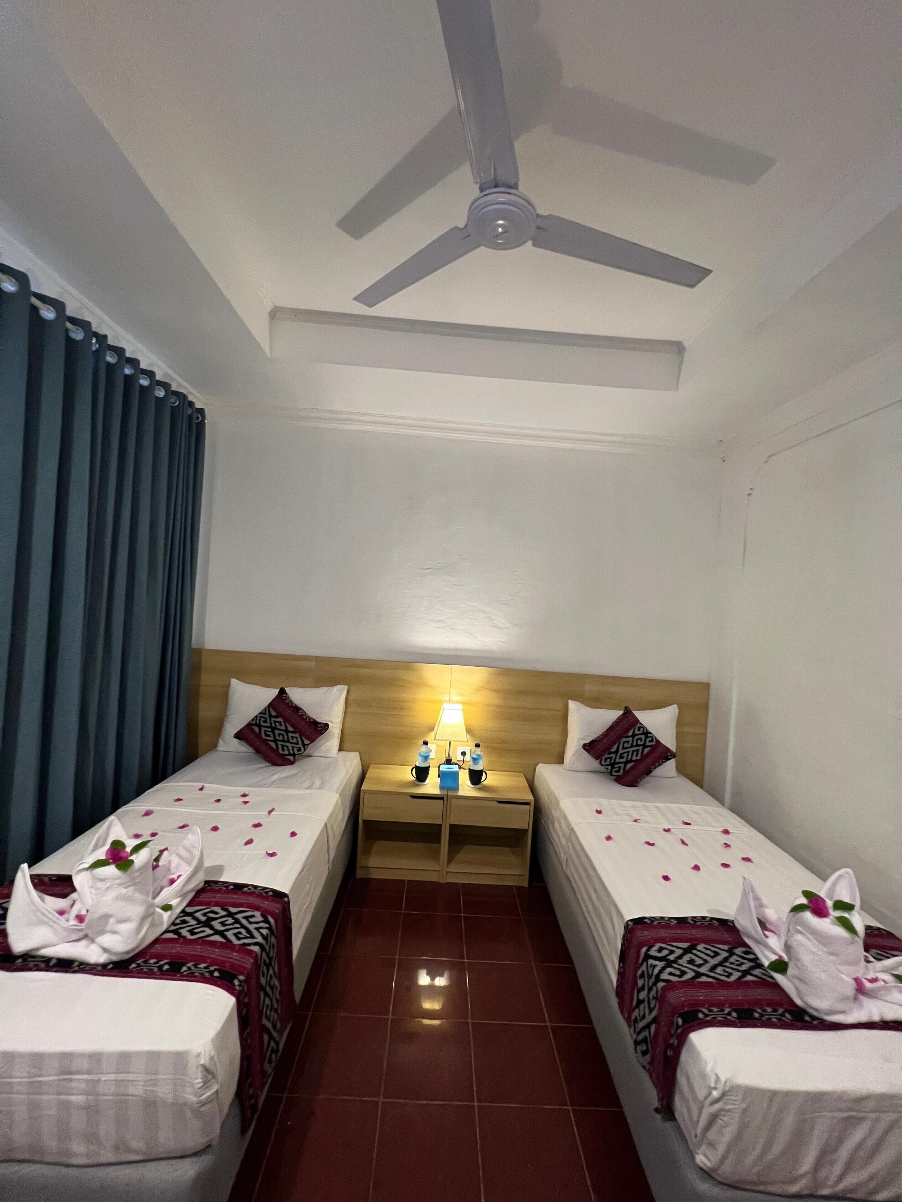 A hotel room with two single beds, a wooden headboard, a nightstand with a lamp, and a ceiling fan.